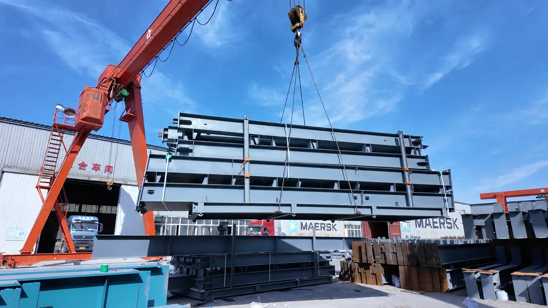 Bundle of fabricated H-section steel members lifted by gantry crane beside export containers